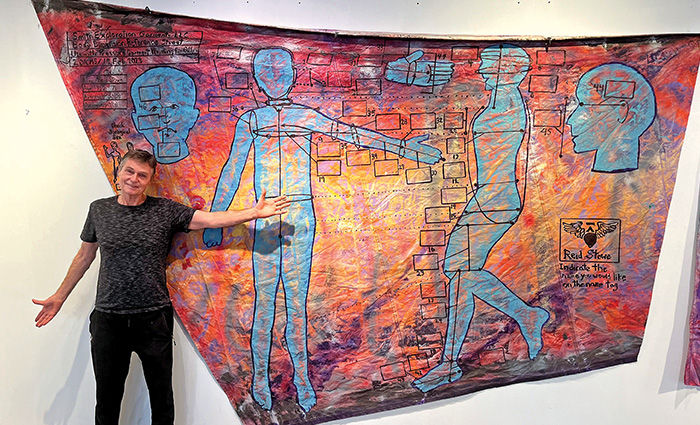 Adult male wearing a dark t-shirt and dark pants posing for a photo in front of a large piece of artwork.