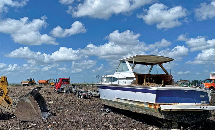 An old and rusted blue and white boat in a landfill with a various trucks and construction equipment.