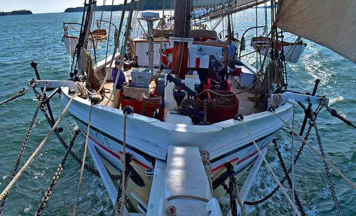 View front the bow of a large white sailboat out on open waters during a sunny day with land in the far background.