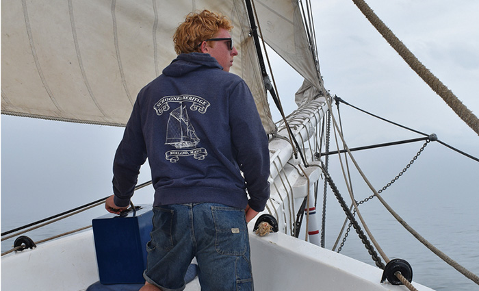 Young red-headed adult male wearing a navy hooded sweatshirt, black sunglasses, jean shorts  and black rain boots looking out from the bow of a white sailboat.