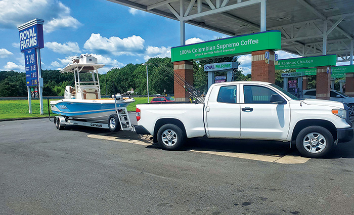 White truck towing a light blue boat  at a roadside filling station on a sunny day.
