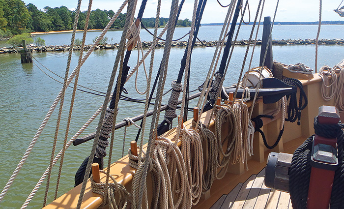 View of numerous tan ropes on the side of a large wooden sailboat out on the water during the day.