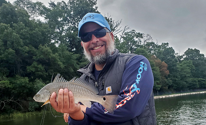 Middle-aged man with a gray beard wearing black sunglasses, a blue ballcap and navy long sleeve shirt proudly displaying a redfish caught.