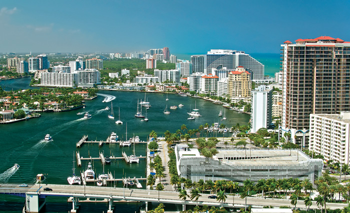 Aerial view of a large marina with various boats docked off the coast of a city with large buildings in the background. 