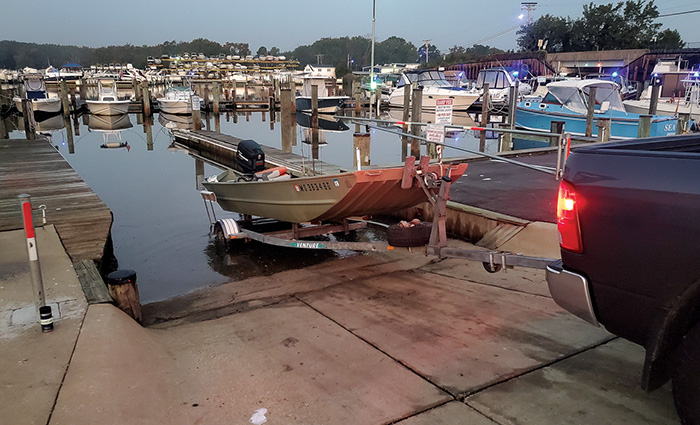 A small silver fishing boat on a trailer being backed in by a black truck to a marina at dusk.