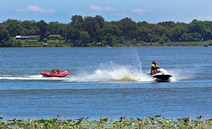 Adult male on a jet ski pulling two young adult females on a red raft out on a large lake.