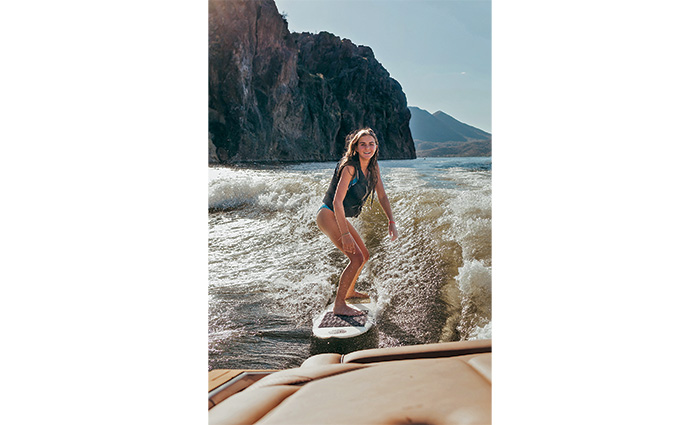 Blonde female wearing a gray life jacket on a white wakeboard on the water.