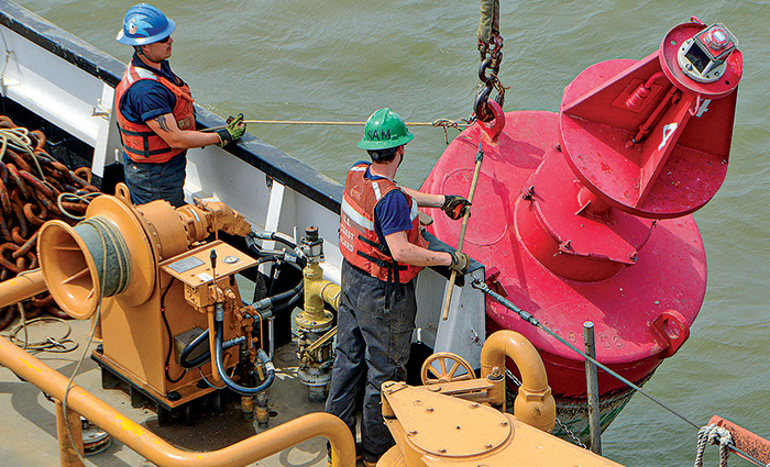 Two men wearing green hard hats and orange life jackets pulling a large red buoy from the water.