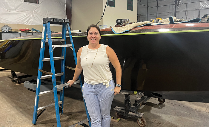 Brunette female wearing a white shirt and blue pants standing next to a ladder and boat inside a warehouse.