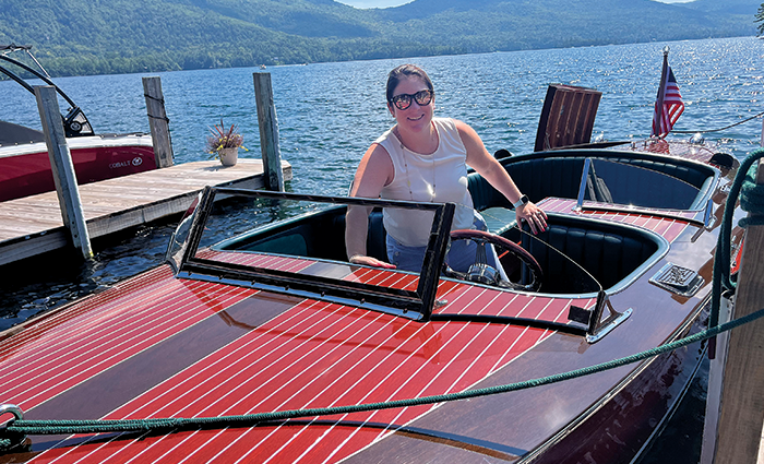 Brunette female wearing sunglasses, a white shirt and blue pants at the wheel of a boat on a dock with clear blue water in the background.