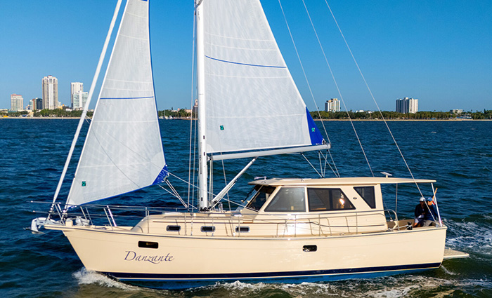 White vessel with blue trim and white sails out on open waters with sunny skies and buildings in the background.