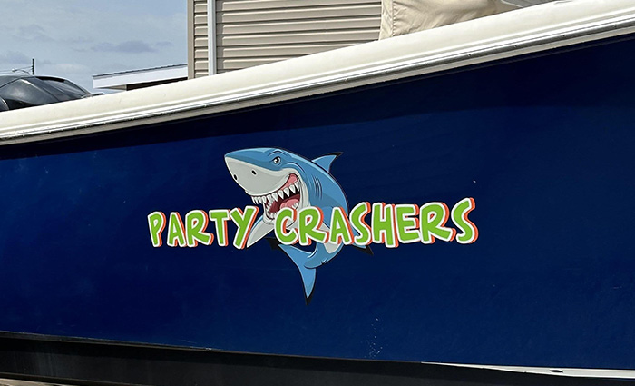 Neon green lettering on the side of a boat reading ' Party Crashers' with a cartoon shark overlayed. 